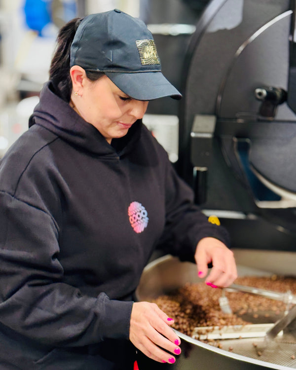 A woman wearing a black hoodie and baseball cap stands beside a large coffee roasting machine, handling coffee beans in a metal tray inside a roastery.